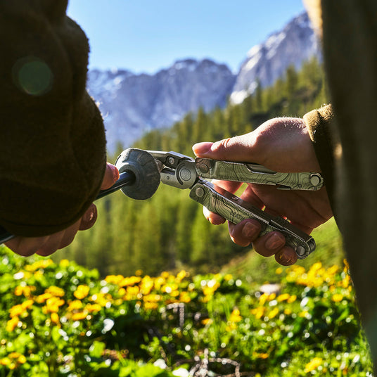 Translation missing: en.Close-up of a person using a Leatherman Rebar multi-tool to adjust or tighten outdoor gear. The scene is set outdoors with mountains, green pine forest, and yellow wildflowers under a clear blue sky