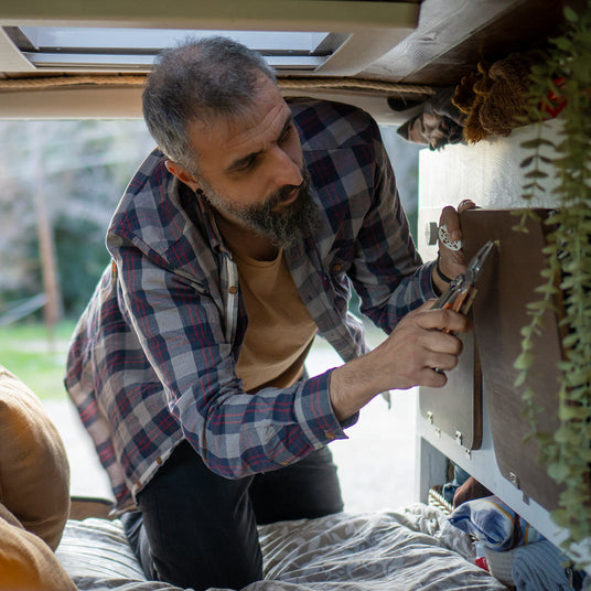 Translation missing: en.Person using a Leatherman Signal multi-tool to make repairs inside a camper van, kneeling on a bed while gripping the tool to tighten a fastener on a cabinet panel