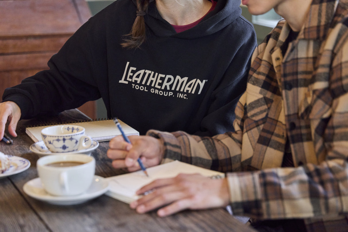 Translation missing: en.Two people sitting at a table with coffee, pastries and notebooks. One is wearing a black Leatherman Blade & Grain hoodie with 'Leatherman Tool Group Inc.' on the front