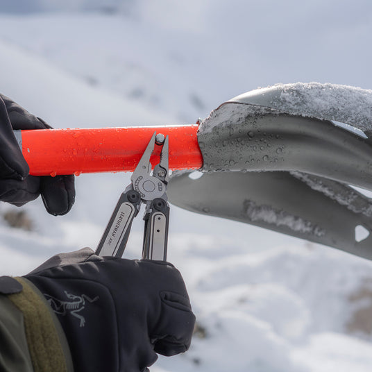 Translation missing: en.Person wearing black gloves using a Leatherman Arc multi-tool to tighten a bolt on an orange-handled snow shovel in a snowy mountain environment
