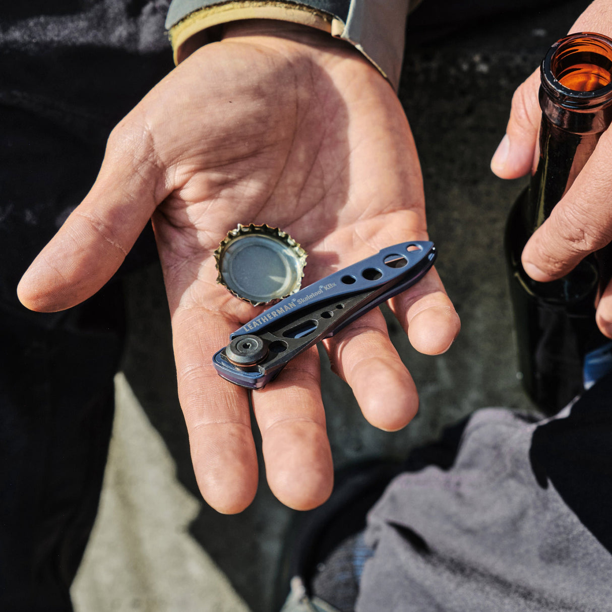 Translation missing: en.Hand holding a blue Skeletool KBX and bottle cap next to a person gripping an open glass bottle