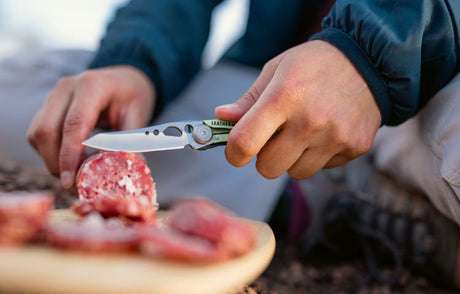 Person slicing salami with a Leatherman Skeletool KB knife on a wooden board outdoors.