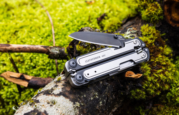 Leatherman multi-tool with knife blade extended, resting on green moss and a tree branch.