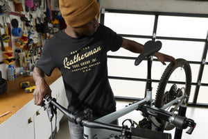 Translation missing: en.A person wearing a black Leatherman Vintage Tee working on a mountain bike in a garage workshop, surrounded by tools and equipment