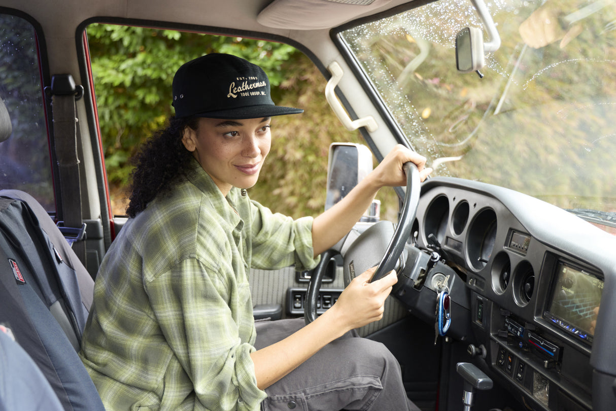 Translation missing: en.A woman wearing a black Leatherman Vintage 5-Panel Hat sitting in the driver’s seat of a vehicle, smiling while holding the steering wheel