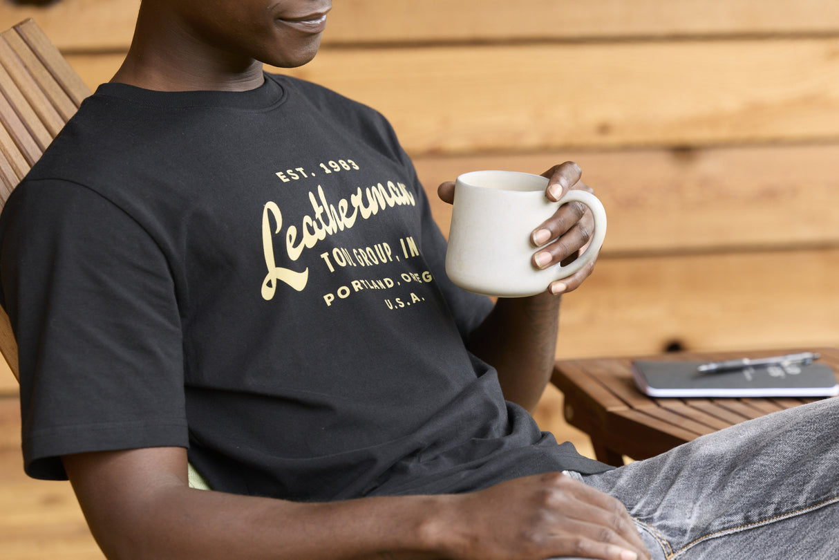 Translation missing: en.A person wearing a black Leatherman Vintage Tee relaxing on a wooden deck chair, holding a white mug with a notebook and pen on a small table nearby