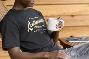 Translation missing: en.A person wearing a black Leatherman Vintage Tee relaxing on a wooden deck chair, holding a white mug with a notebook and pen on a small table nearby