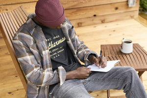 Translation missing: en.A person wearing a black Leatherman Vintage Tee sitting on a wooden chair outdoors, writing in a notebook with a mug of coffee on a nearby table