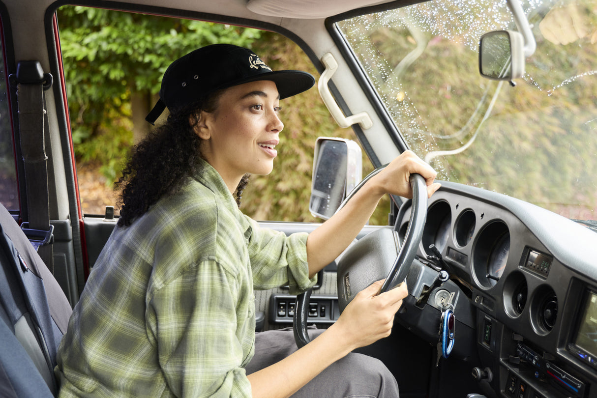 Translation missing: en.A woman wearing a black Leatherman Vintage 5-Panel Hat driving a vehicle with morning dew on the windshield, smiling