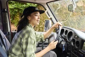 Translation missing: en.A woman wearing a black Leatherman Vintage 5-Panel Hat driving a vehicle with morning dew on the windshield, smiling