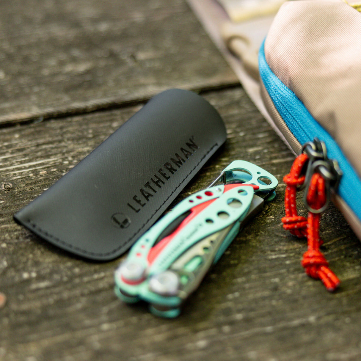 Translation missing: en.A black Leatherman Hypalon tool sleeve placed on a wooden surface next to a folded Paradise-colored Skeletool® CX multi-tool, with a beige and blue-zippered pouch partially visible in the background