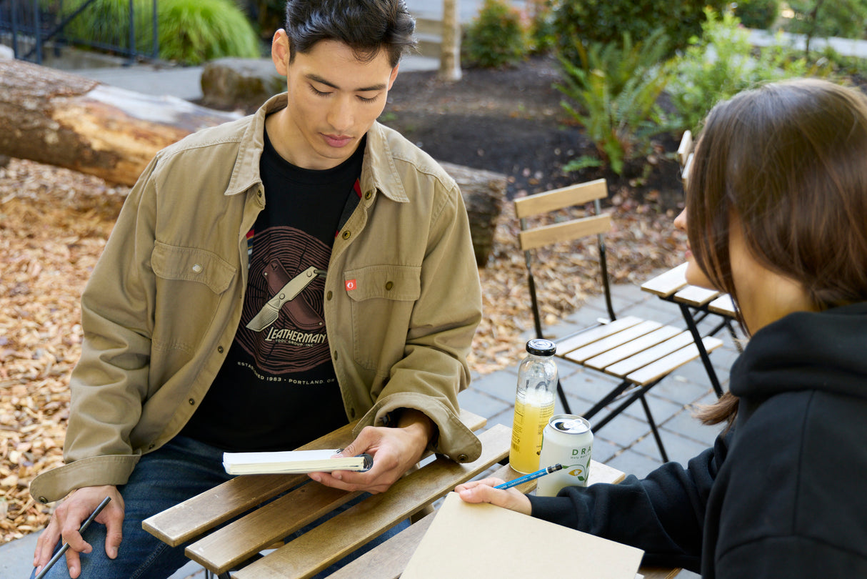 Translation missing: en.A man sitting at a table wearing a black Leatherman Blade &amp; Grain tee with a woman wearing a black hoodie, both equipped with a notebook and pencil