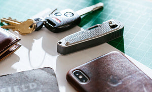 Translation missing: en.Brown Leatherman Wallet, car keys, Leatherman T2, gray note pad, and Iphone on top of white piece of paper placed on green grid table