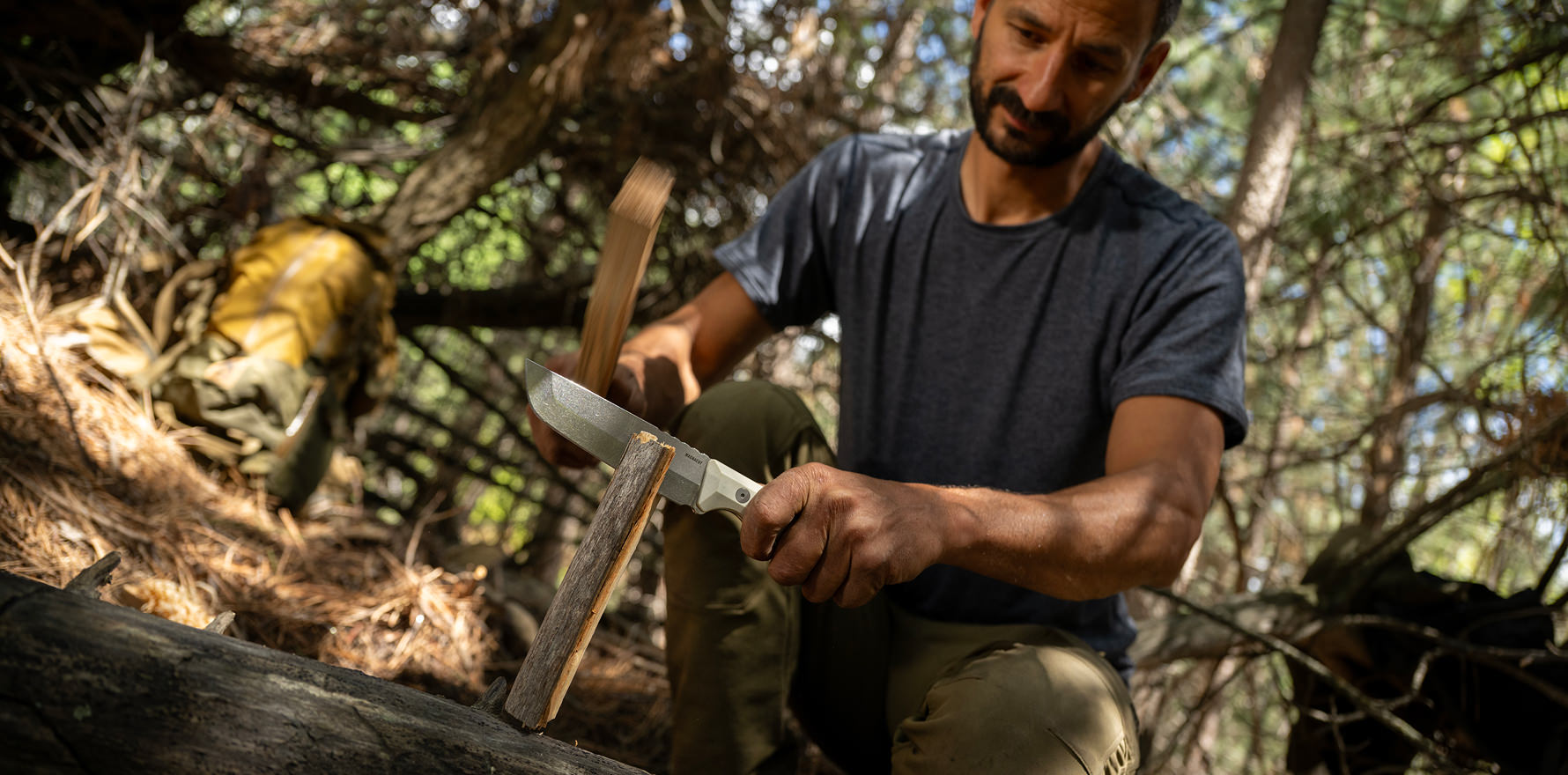 A man is using a Leatherman Pioneer knife to cut wood in a forest setting, surrounded by trees and natural scenery.