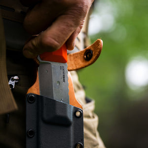 Translation missing: en.A person gripping a Leatherman Orange pop Pioneer, partially sheathed in a leather cover, showcasing the blade's sharp edge and craftsmanship