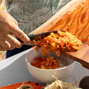 Translation missing: ko.Person using a Leatherman Rustle Evergreen knife with a green handle to transfer freshly chopped tomatoes and vegetables from a wooden cutting board into a white bowl, outdoors on a sunny day