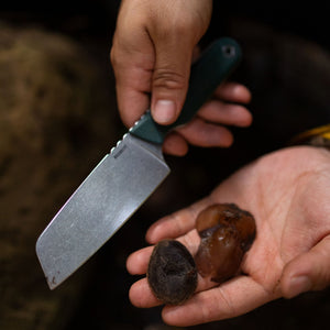 Translation missing: ko.Person holding a Leatherman Rustle Evergreen knife with a green handle in one hand and dried dates in the other, suggesting outdoor food preparation or foraging