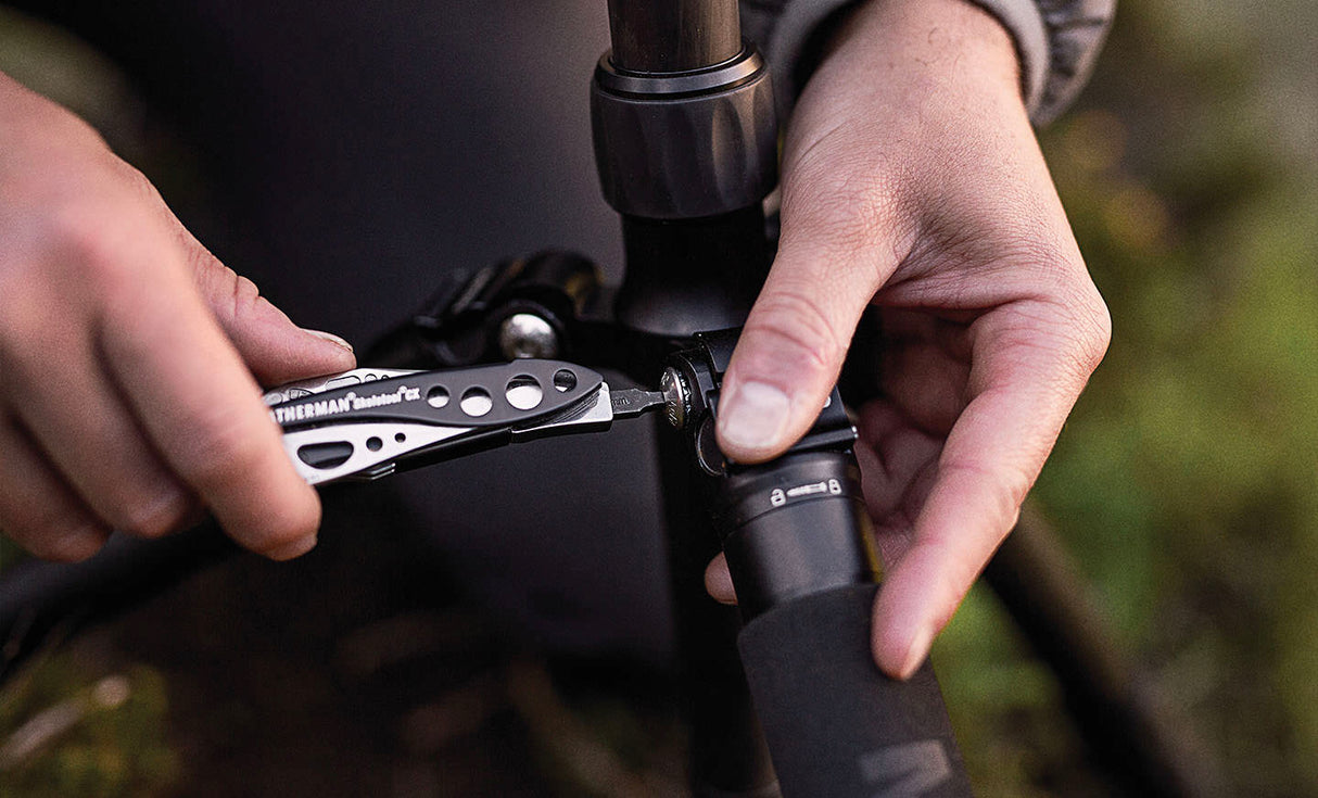 Translation missing: en.Person using a Black &amp; Stainless Leatherman Skeletool® CX multi-tool to adjust a camera tripod in an outdoor setting