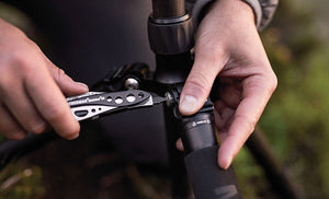 Translation missing: en.Person using a Black &amp; Stainless Leatherman Skeletool® CX multi-tool to adjust a camera tripod in an outdoor setting