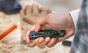 Translation missing: en.Person holding a closed OD Green Leatherman Skeletool® CX multitool over a wooden work bench