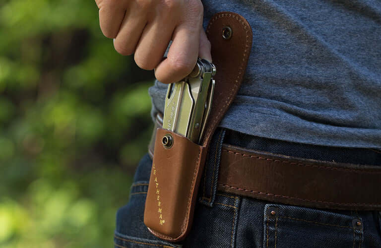 A person reaching for a Stainless Steel Leatherman Wave® Plus multi-tool in a brown leather sheath on their belt loop