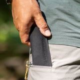 Translation missing: en.A person placing a black leather Leatherman tool sleeve, with a tool inside, into the side pocket of light gray pants; the sleeve features an embossed vertical "LEATHERMAN" logo and circular emblem